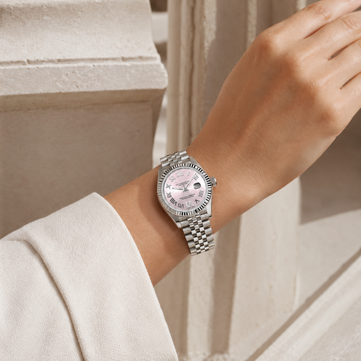 A silver-tone wristwatch with a pink dial and matching pink hour markers, worn on a model's wrist. The watch has a silver-tone metal bracelet and is photographed outdoors against a light beige stone wall.