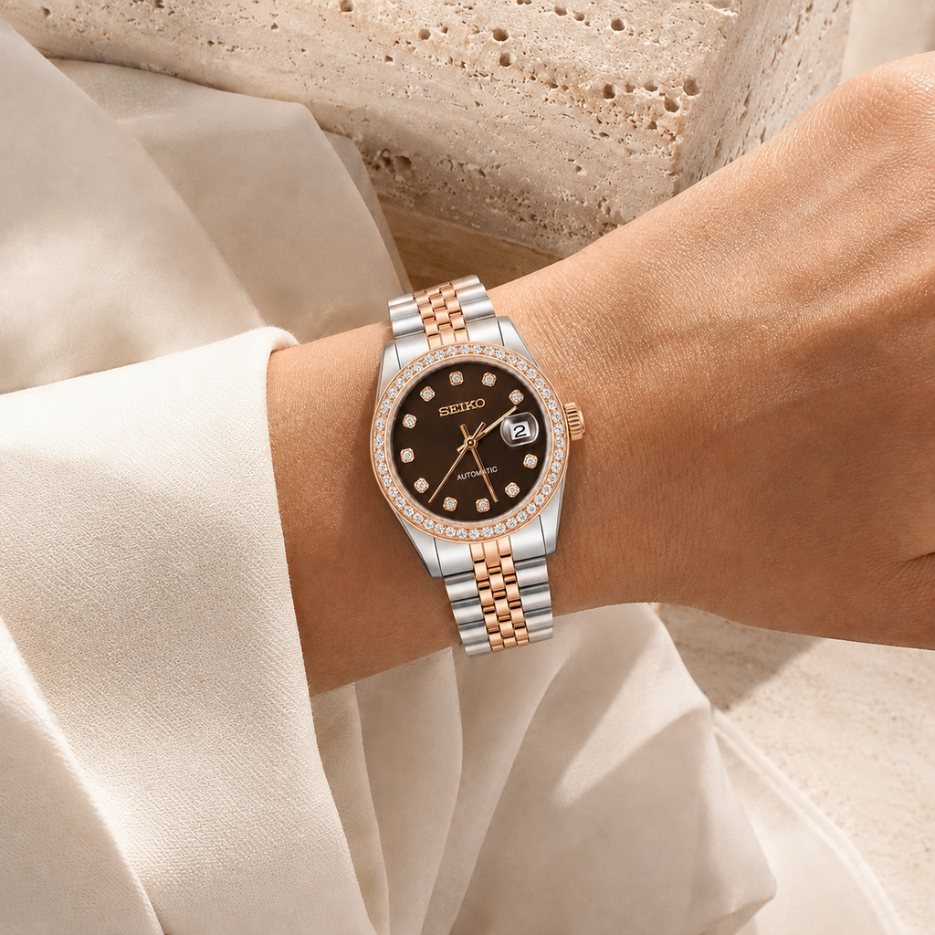 A wristwatch with a two-tone silver and rose gold metal bracelet, a black dial, and a crystal-studded bezel, shown worn on a person's wrist.