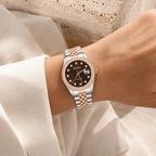 A wristwatch with a two-tone silver and rose gold metal bracelet, a black dial, and a crystal-studded bezel, shown worn on a person's wrist.