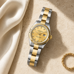 A two-tone stainless steel and gold wristwatch with a gold dial and fluted bezel, styled on a textured beige surface next to draped cream satin fabric and a gold chain bracelet.