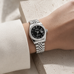 A silver stainless steel wristwatch with a black dial and fluted bezel, shown worn on a person's wrist against a light stone and white fabric background.
