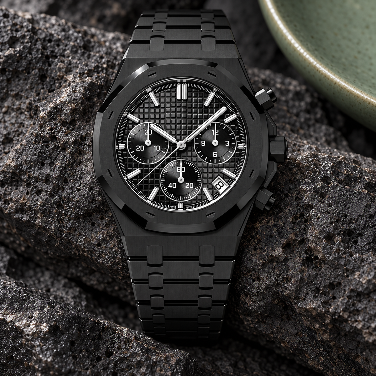 Black Phantom chronograph laid on volcanic stone beside sage ceramic