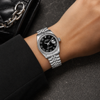 A silver-tone stainless steel wristwatch with a black dial and fluted bezel, shown worn on a person's wrist. The watch has Roman numeral hour markers, a date window at the 3 o'clock position, and a matching steel bracelet.