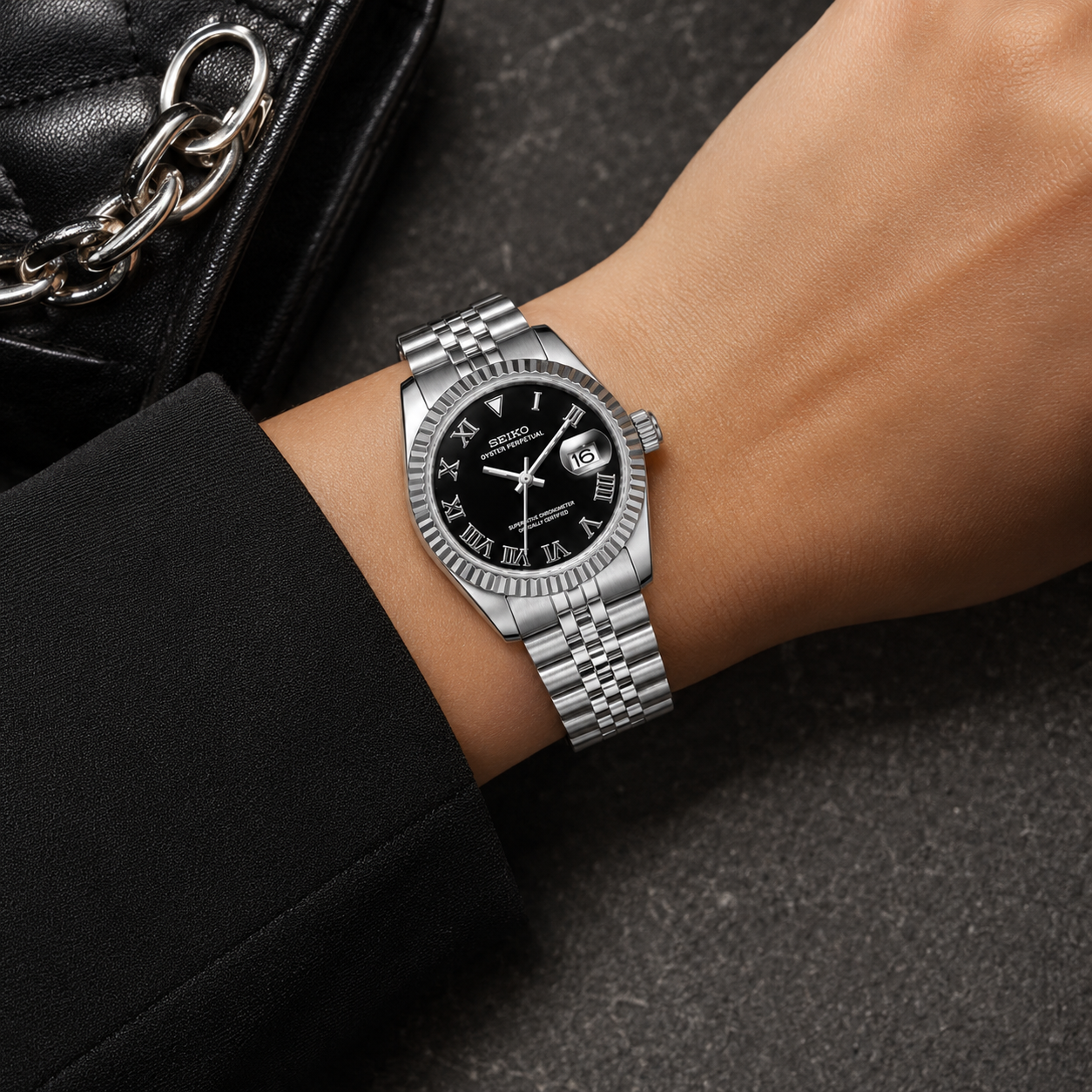 A silver-tone stainless steel wristwatch with a black dial and fluted bezel, shown worn on a person's wrist. The watch has Roman numeral hour markers, a date window at the 3 o'clock position, and a matching steel bracelet.